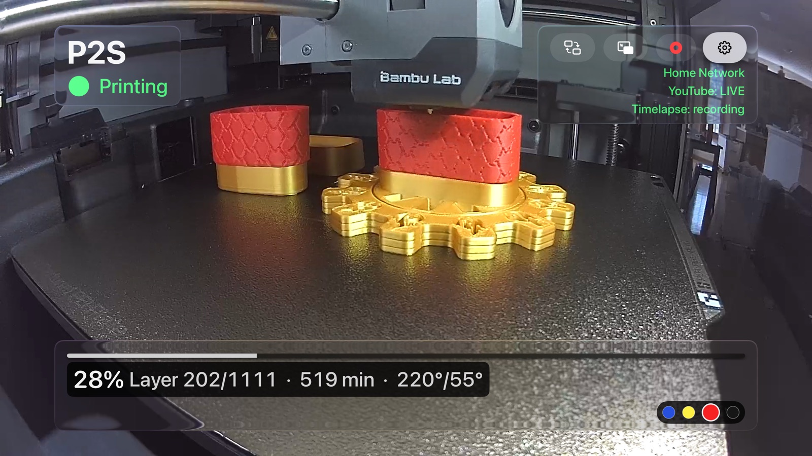 AmbientCast on an Apple TV: the printer's live camera feed fills the screen, with the printer name and Printing status in the top corner, a status bar reading 28 percent, layer 202 of 1111, 519 minutes and 220 over 55 degrees, and the AMS filament colours beside it.